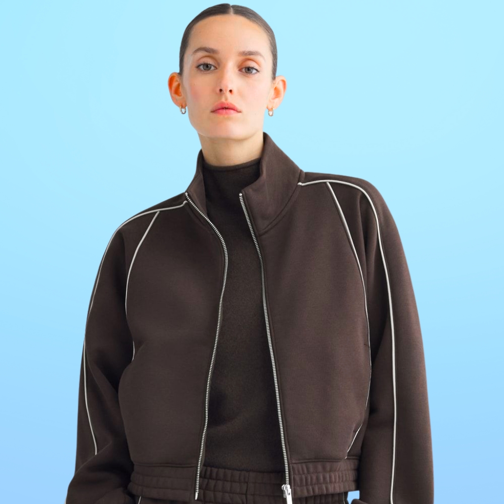 Person wearing a brown zip-up jacket with white seams over a black top, standing against a light blue background.