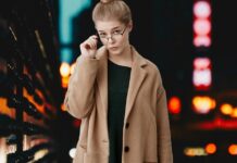 Crombie Coat Explained: History, Fit, Fabric, and Styling A stylish woman in a camel coat and dark dress, wearing glasses, stands against a dimly lit urban backdrop with blurred city lights.