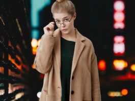 A stylish woman in a camel coat and dark dress, wearing glasses, stands against a dimly lit urban backdrop with blurred city lights.