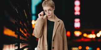 Crombie Coat Explained: History, Fit, Fabric, and Styling A stylish woman in a camel coat and dark dress, wearing glasses, stands against a dimly lit urban backdrop with blurred city lights.
