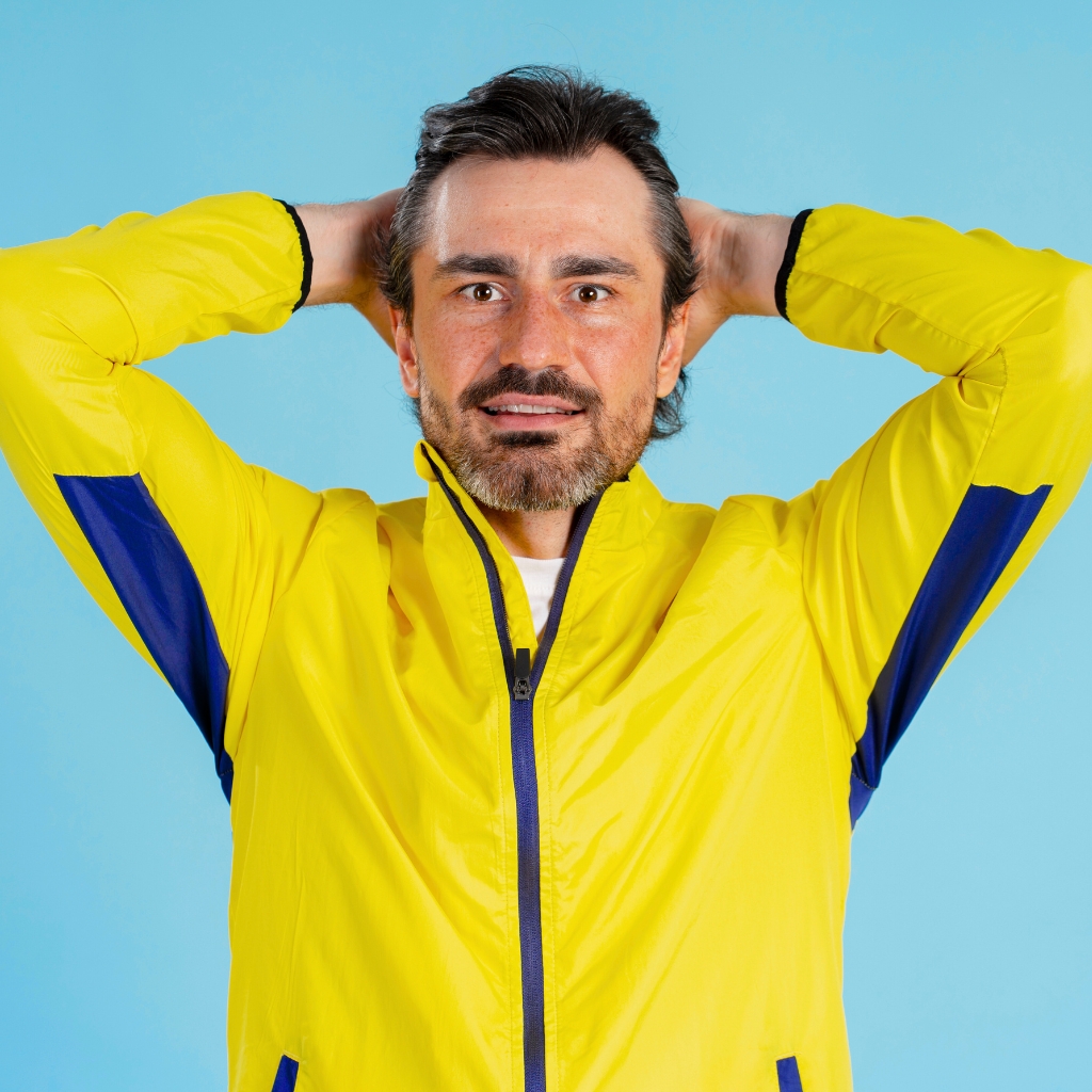 A man with a trimmed beard wearing a bright yellow windbreaker with blue accents, standing against a light blue background. He has his hands behind his head and looks directly at the camera with a surprised or amused expression.