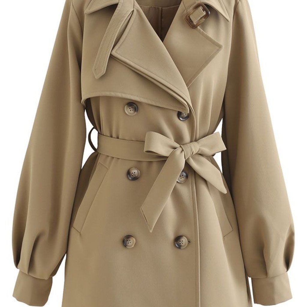 A close-up view of a beige trench coat with a double-breasted front, wide lapels, a belted waist tied in a bow, and puffed sleeves. The coat features large buttons and a decorative shoulder strap.