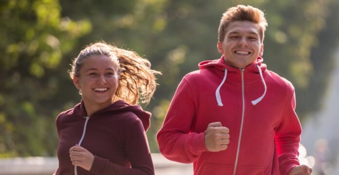 Two young people jogging outdoors on a sunny day, woman in a maroon hoodie on the left and man in a bright pink hoodie on the right, both smiling.