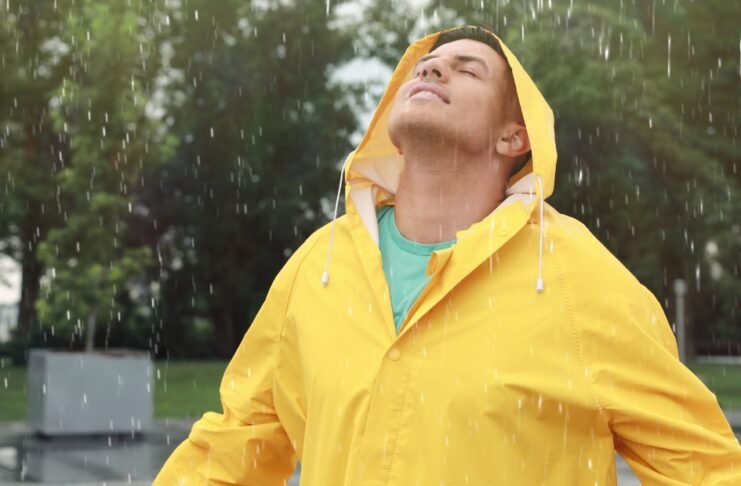A man wearing a bright yellow raincoat stands with his face tilted upward in light rain, droplets falling around him in a park setting.