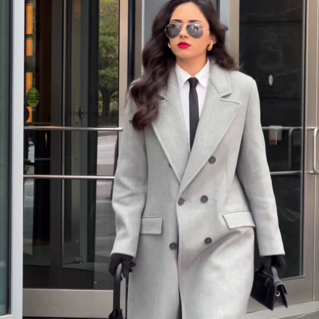 A stylish woman walking outdoors in a light gray oversized double-breasted wool coat, white shirt, black tie, black gloves, black handbag, and aviator sunglasses. She has long wavy dark hair and bright red lipstick.