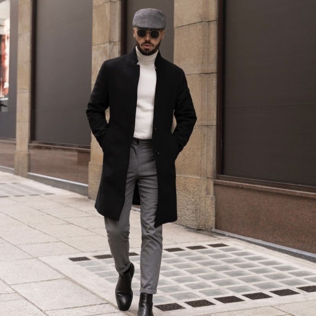 A man walking on a city sidewalk wearing a black long coat, white turtleneck, gray trousers, black ankle boots, a gray flat cap, and round sunglasses.
