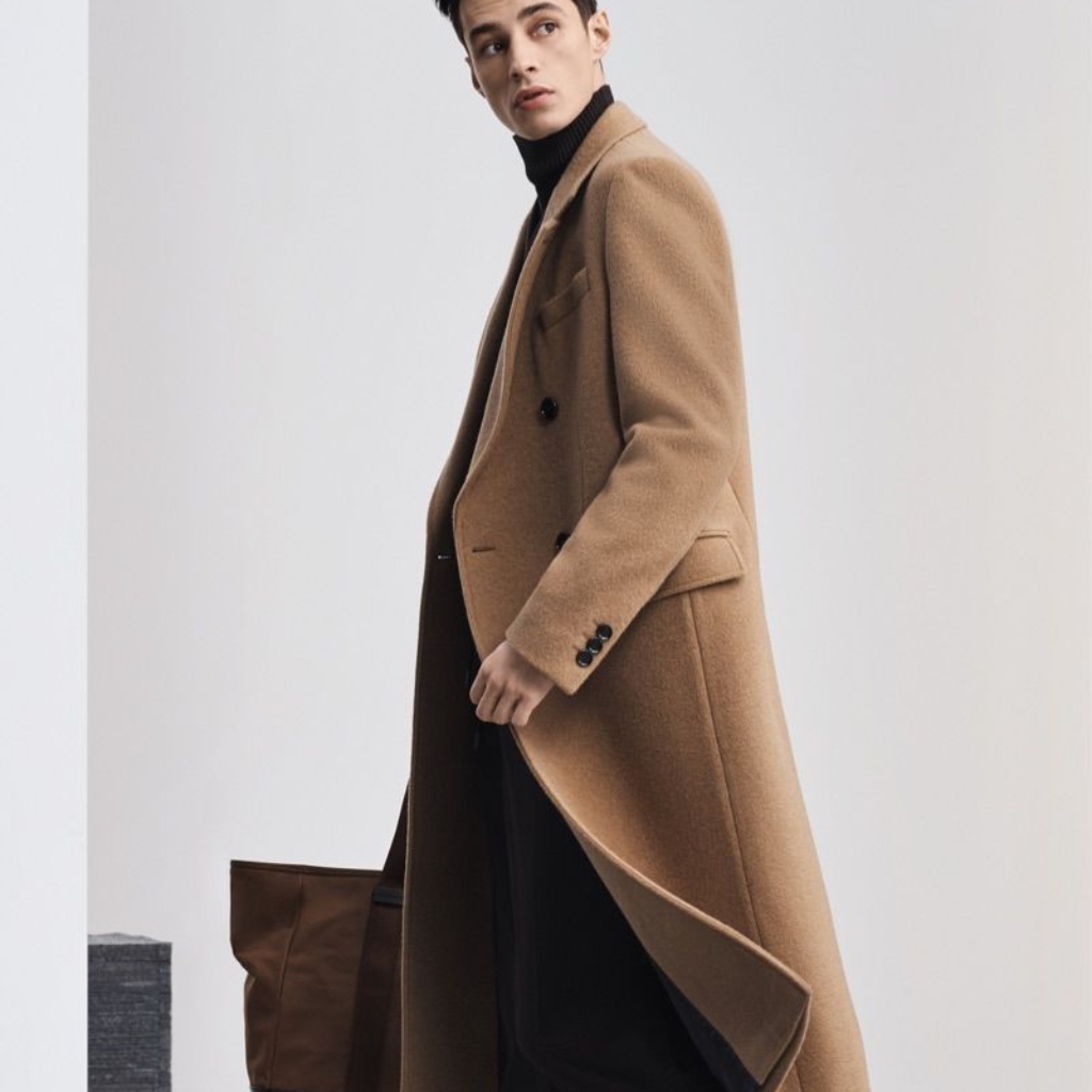 A man wearing a long camel coat over a black turtleneck, standing indoors with a neutral background.