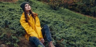 Insulated Rain Jacket: Warm, Waterproof Protection Guide A woman with a black beanie sits on a hillside covered in low, dense green shrubs. She wears a bright yellow rain jacket, dark jeans, and brown lace-up boots, with her eyes closed and a relaxed expression.