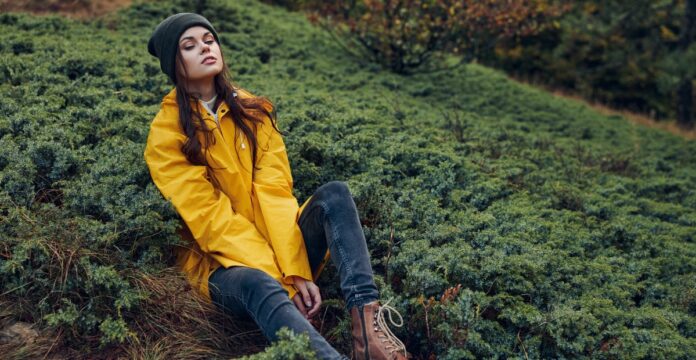 A woman with a black beanie sits on a hillside covered in low, dense green shrubs. She wears a bright yellow rain jacket, dark jeans, and brown lace-up boots, with her eyes closed and a relaxed expression.