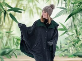 A woman wearing a knitted hat and a dark oversized poncho stands on a wooden path surrounded by tall green bamboo plants, smiling and looking to the side.