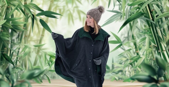 A woman wearing a knitted hat and a dark oversized poncho stands on a wooden path surrounded by tall green bamboo plants, smiling and looking to the side.