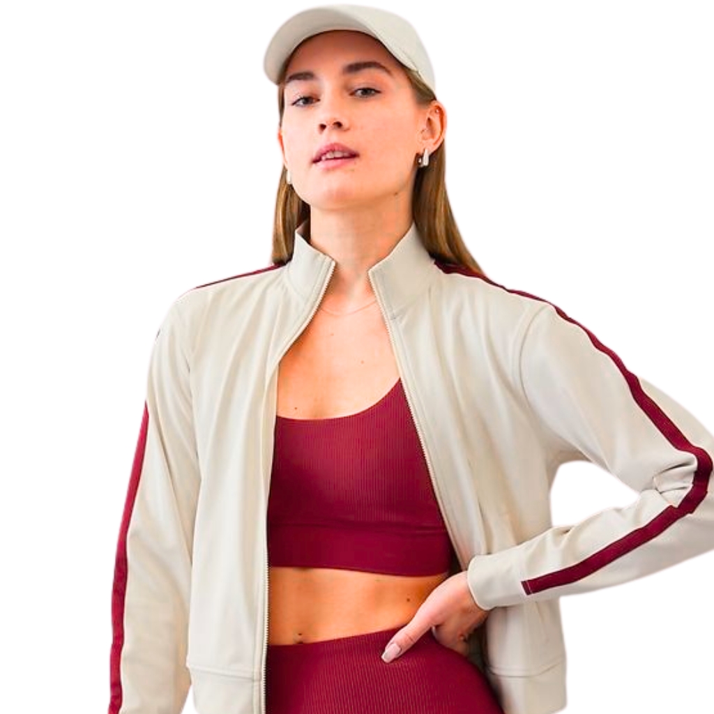 A fashion model wearing a light beige zip-up jacket with burgundy side stripes over a matching burgundy sports bra and high-waisted leggings, plus a light-colored cap. She stands with one hand on her hip, facing slightly to the right.