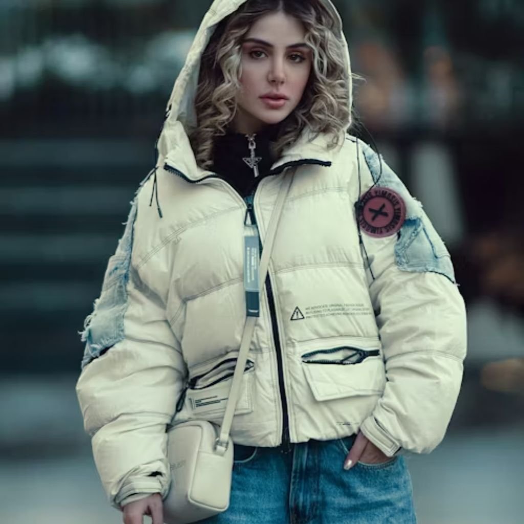 A woman wearing a voluminous cream-colored puffer jacket with blue denim jeans and a small white crossbody bag. She has wavy blonde hair under a hood and looks off to the side.
Daily Wear Winter Jacket: