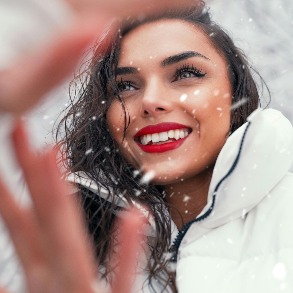 A smiling woman with red lipstick and wet hair leaning toward the camera in a snowy scene, wearing a white puffer jacket.Minimalist Winter Jackets: