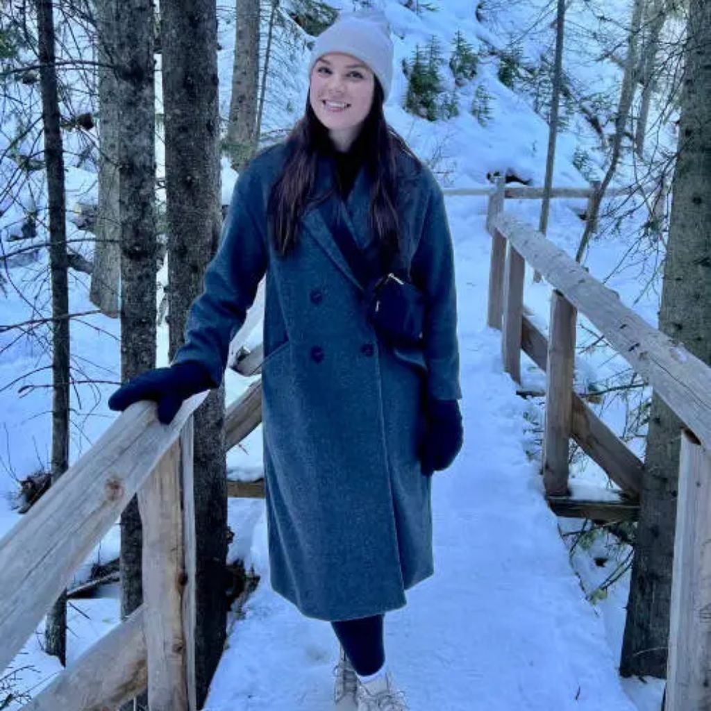 Sustainable Wool Winter Coat: Warmth with a Green Touch Eco 9 Alt text: A woman stands on a snow-covered wooden bridge wearing a blue-grey knee-length coat, a light grey beanie, dark gloves, and a small black purse. Snowy trees surround the scene.
Sustainable Wool Winter Coat: