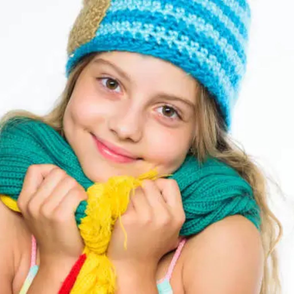 A smiling young girl wearing a blue knit beanie and a teal scarf, holding the scarf close to her chin with both hands.Warmest Fabrics for Winter Clothing:
