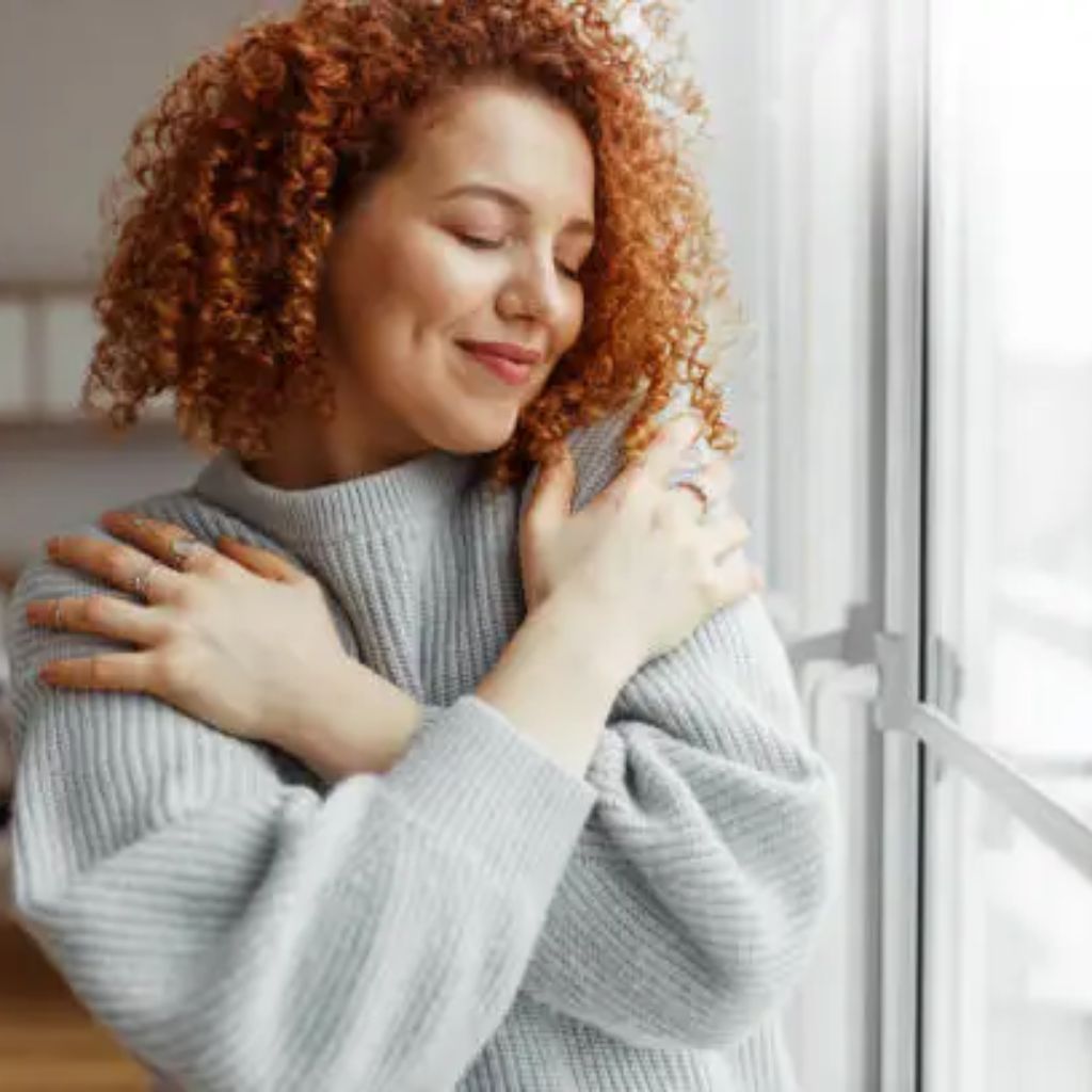 Soft Winter Fabrics for Sensitive Skin: Cozy & Itch-Free Now 10 A woman with curly red hair wearing a light gray knit sweater, standing by a window with her arms crossed over her chest and a gentle smile.Soft Winter Fabrics for Sensitive Skin: