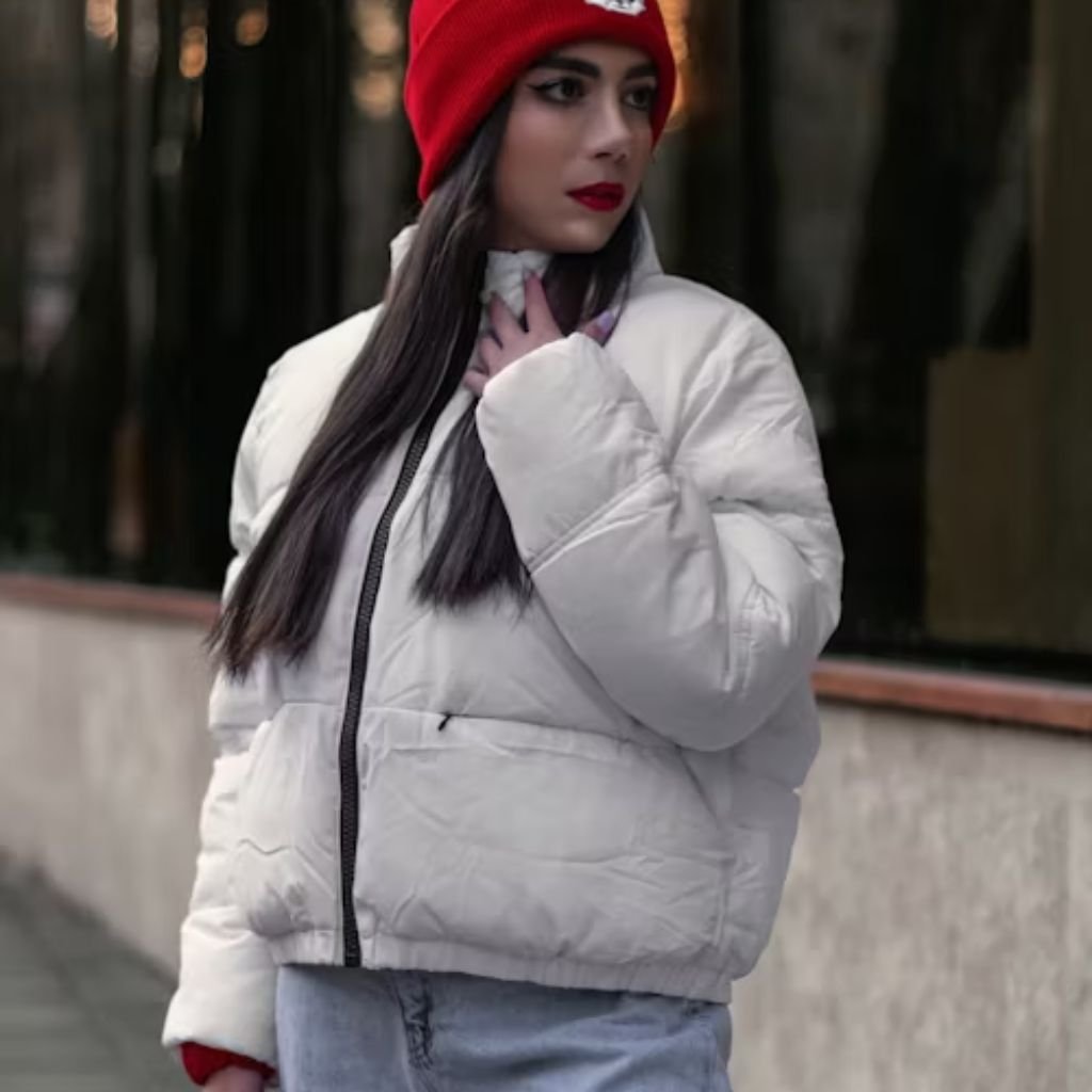 A woman wearing a white puffy winter jacket and a bright red beanie stands outdoors on a city sidewalk. She has long dark hair and bold red lipstick, and she is touching the collar of her jacket with one hand.
Daily Wear Winter Jacket: