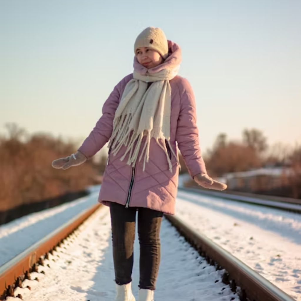 A woman stands on snow-covered train tracks in winter, wearing a pink quilted coat, white boots, a cream hat and scarf, with gloved hands and a scarf wrapped around her neck.
Cozy Casual Winter Jacket:

