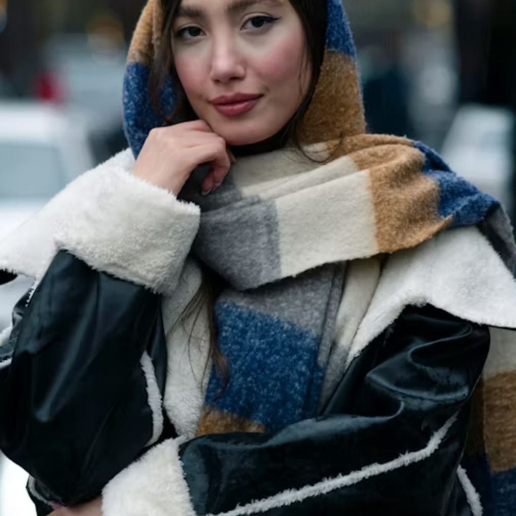 Alt text: A stylish woman wearing a large color-block fleece-and-leather coat and a blue, tan, and white scarf. She rests her chin on her hand and looks at the camera with a soft smile.

Ethical Winter Outerwear Coat: