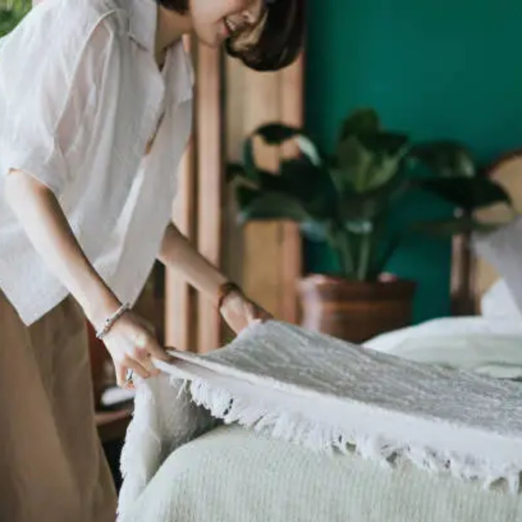 A person in a white shirt lifts and smooths a large white fabric or blanket over a bed, with a soft, textured surface visible in the foreground.Blankets and Bedding fabic
