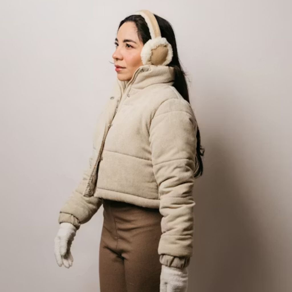 A woman stands in profile wearing a cream-colored puffer jacket with a high collar, matching mittens, and cream earmuffs. She gazes to the left, with long dark hair visible beneath the hood.Daily Wear Winter Jacket:
