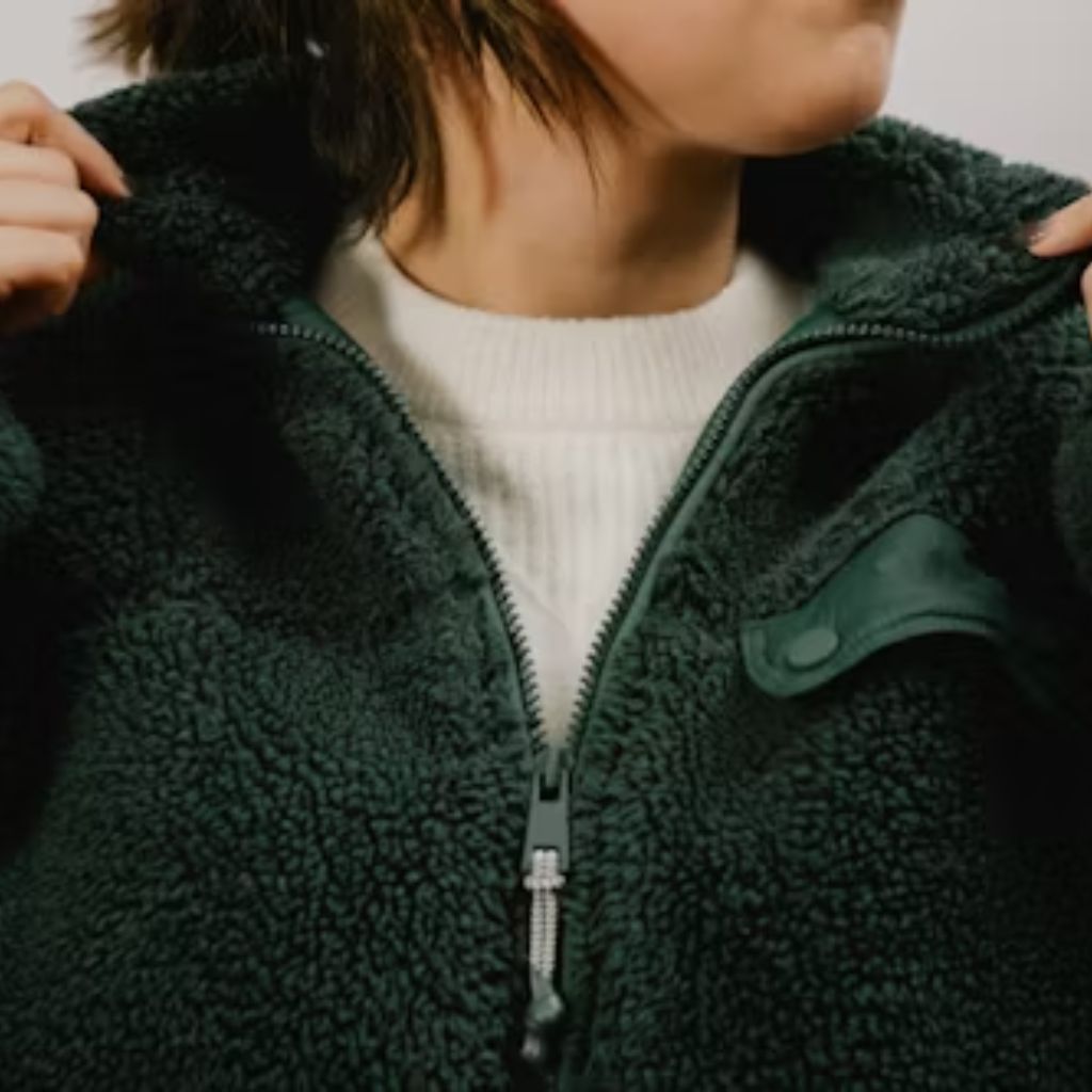 A close-up of a person wearing a dark green fuzzy fleece jacket over a white sweater. The jacket is zipped up with a raised collar, and the person is holding the collar with both hands, highlighting the texture and zipper details.
Cozy Casual Winter Jacket:
