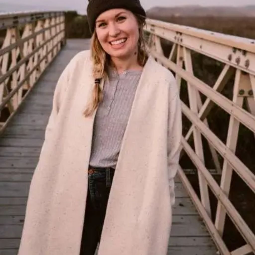 Sustainable Wool Winter Coat: Warmth with a Green Touch Eco 12 Alt text: A smiling young woman with blonde hair stands on a wooden bridge. She wears a dark knit beanie, a light gray knit top, a long light-colored cardigan, and dark jeans. The bridge has pale railings and a wooden deck, with a muted landscape and hints of water in the background.
Sustainable Wool Winter Coat: