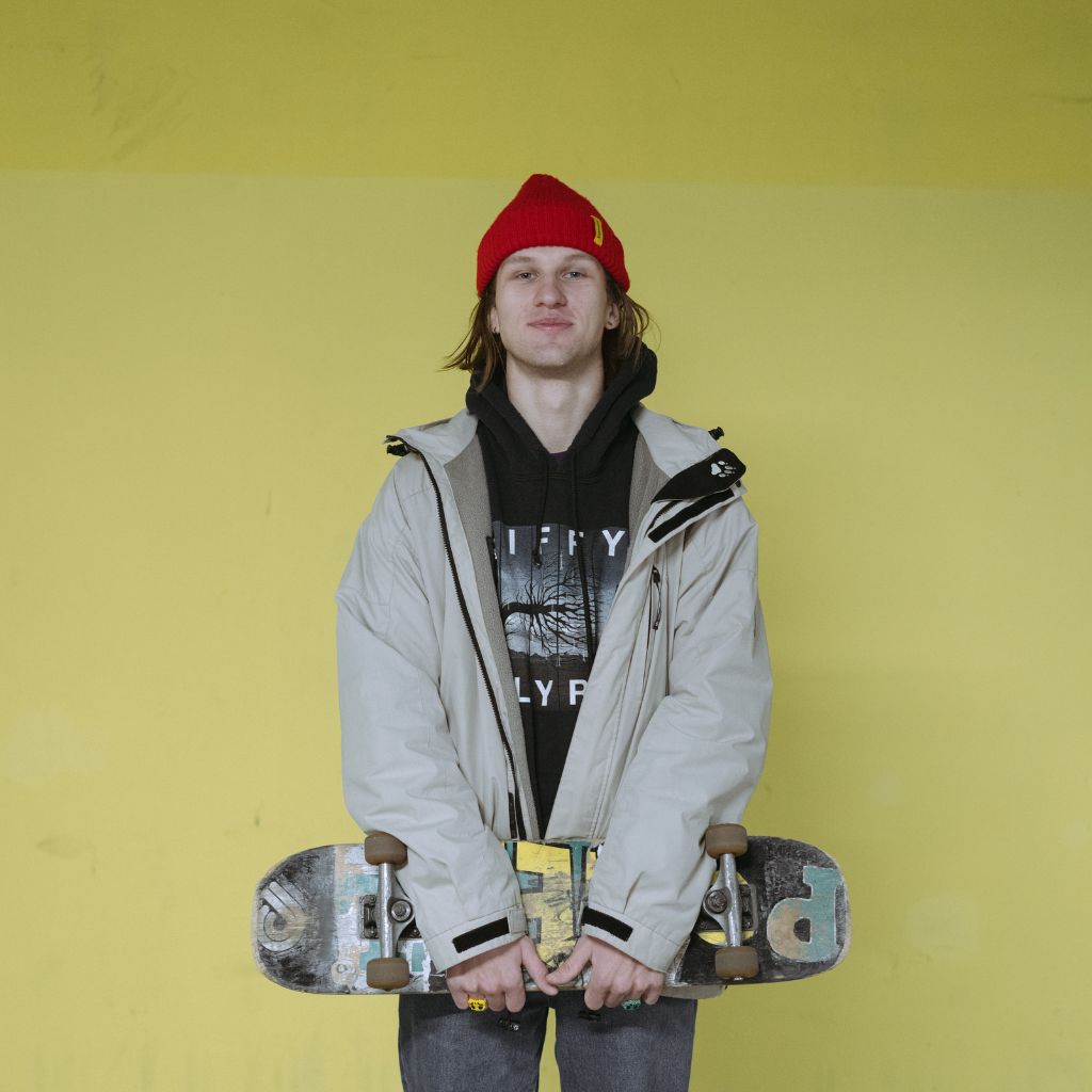 A young man wearing a red beanie, black hoodie, and light jacket stands against a yellow wall, holding a skateboard in front of him.Minimalist Winter Jackets: