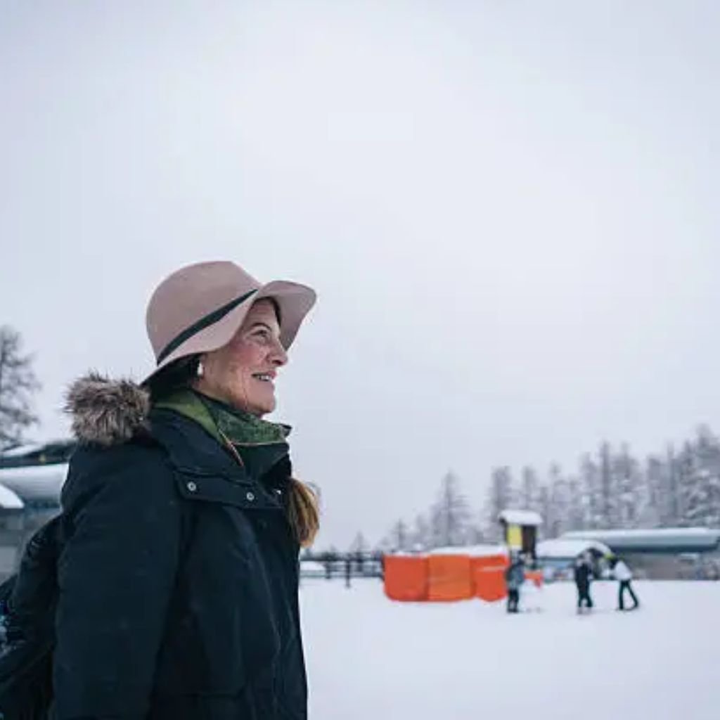 A smiling woman in a winter coat and wide-brimmed beige hat stands outdoors in a snowy landscape, with orange barriers and people in the background.
Sustainable Winter Fabrics for 2025: