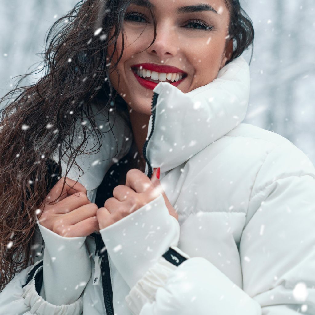 A smiling woman with long hair wearing a white puffer jacket in a snowy scene, holding her jacket closed.Minimalist Winter Jackets: