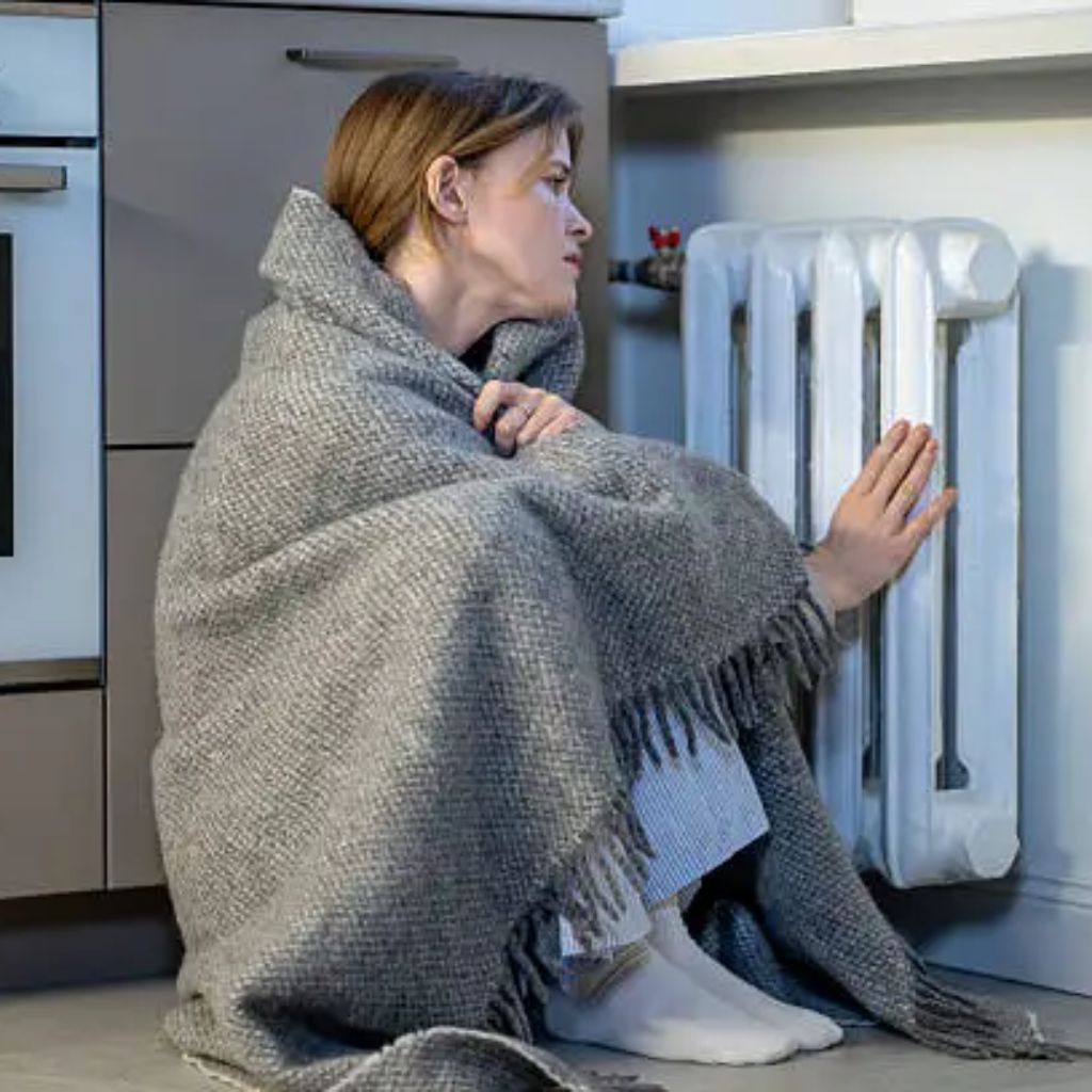 “Woman sitting on the floor by a radiator, wrapped in a thick gray woven blanket with fringe, appearing cold.”Wool Thermal Fabric Explained: