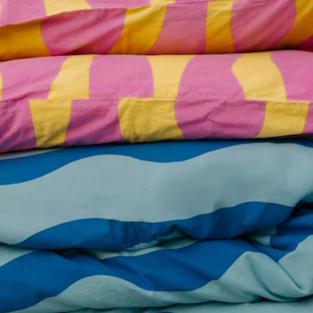 Close-up of stacked colorful bedding: pink and yellow striped blanket on top, followed by a blue and turquoise wave-patterned comforter.Blankets and Bedding fabic
