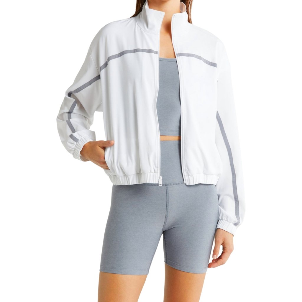 Woman wearing a white zip-up athletic jacket with gray stripes over a light blue sports bra and matching gray bike shorts.