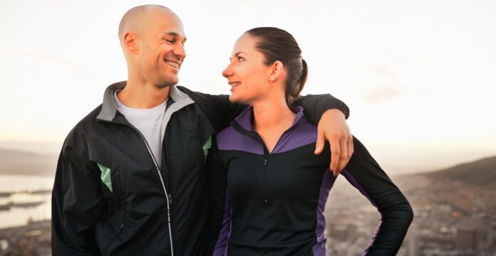 A smiling couple stands close together outdoors with their arms around each other, wearing athletic jackets, overlooking a valley or cityscape at sunset. Minimalist Track Jacket