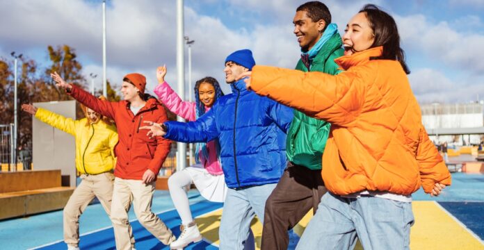 A diverse group of six people wearing colorful jackets—yellow, red, blue, green, orange—stand on a bright outdoor track, smiling and dancing with arms extended. Modern Track Jacket