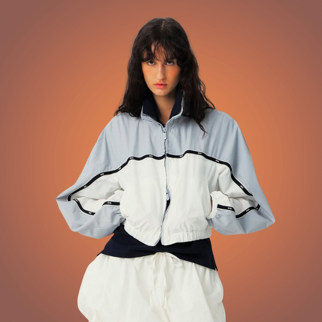 Person with dark hair wearing a light blue and white color-block windbreaker with a black zipper and black detailing, standing against a warm, orange-brown gradient background.