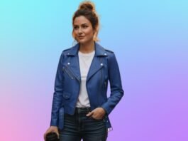 A woman stands wearing a blue navy leather jacket over a white top, with her hair tied up in a bun, against a soft gradient pastel background.