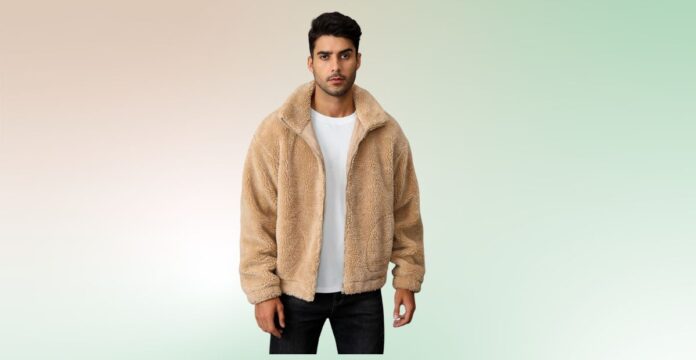 A man wearing a beige plush teddy jacket over a white T-shirt and black jeans stands against a soft gradient background.