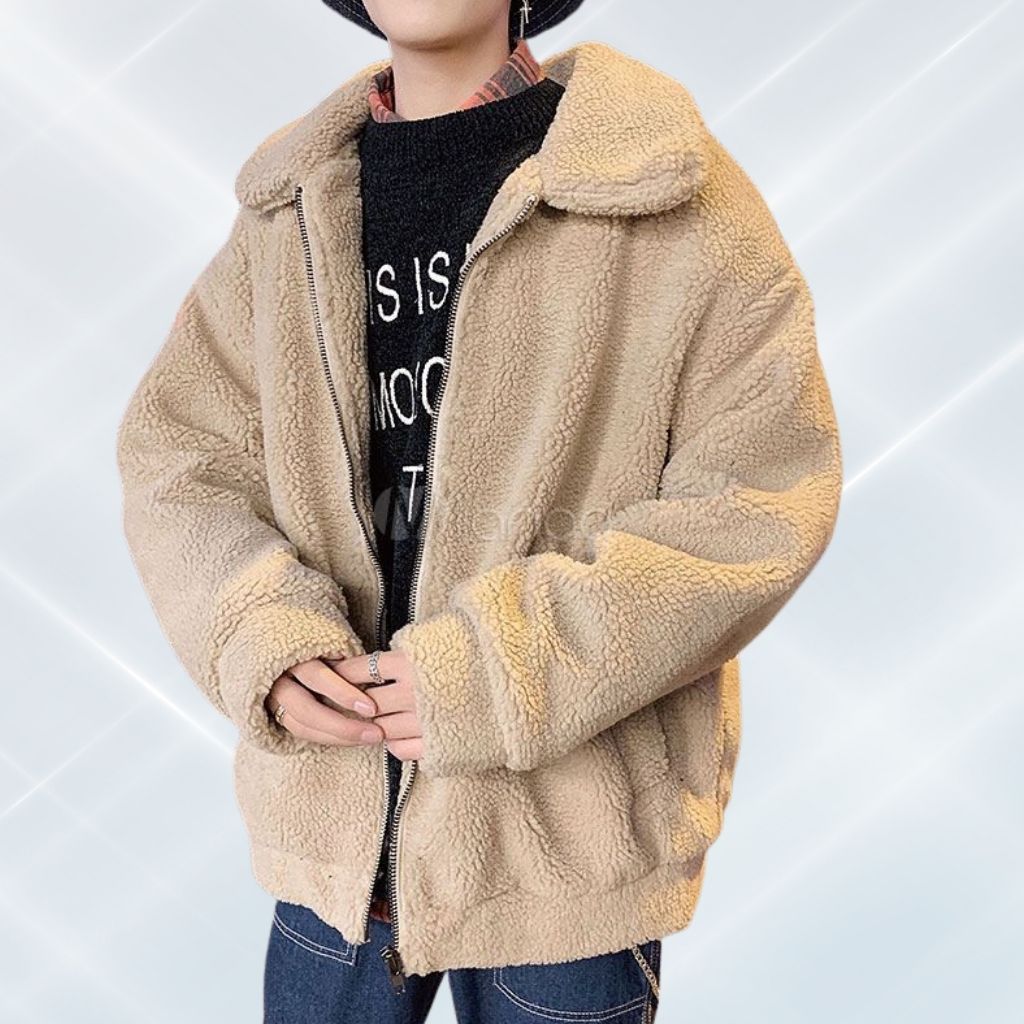 A person wearing a tan, oversized teddy bear-style plush jacket with a front zipper, paired with a black graphic sweatshirt and jeans.
