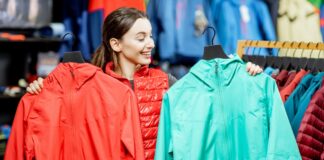 Poncho Rain Jackets: Lightweight Protection for Wet Weather A woman in a red puffy vest stands in a clothing store, holding up a bright red jacket in her left hand and a bright teal jacket in her right hand. Racks of colorful jackets and vests line the background.