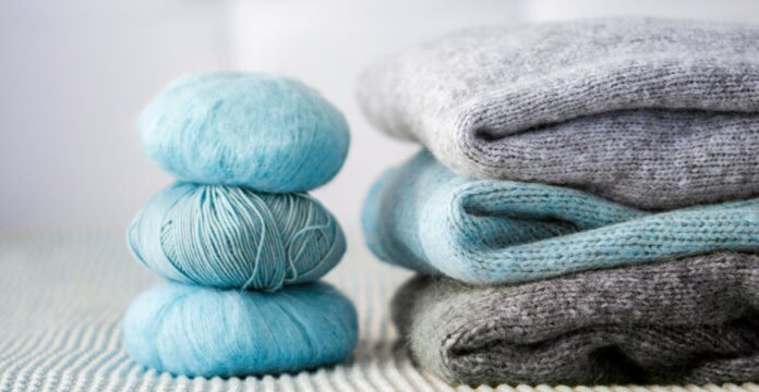 Pure Grade A Cashmere Wool Fabric A stack of folded knit sweaters in gray and turquoise tones with balls of light blue yarn beside them.