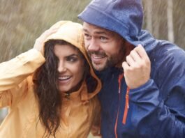 A smiling couple standing in the rain, both wearing rain jackets with hoods up; the woman in a yellow jacket and the man in a blue jacket lean close together, enjoying the moment despite the weather.