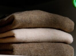 Recycled Cashmere Wool Fabric A stack of folded sweaters in neutral colors (brown, beige, and cream) sits on a shelf, with a wooden basket and a green plant visible on the sides.Recycled Cashmere Wool Fabric
