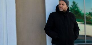 Cozy Casual Winter Jacket: Stay Warm Without Losing Style A man wearing a black hooded winter coat and beanie stands near a wall, looking to the side. He has a beard and a glass window reflecting trees in the background. Cozy Casual Winter Jacket: