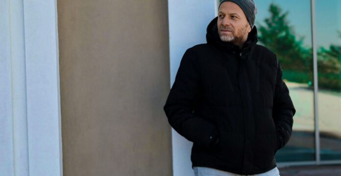 A man wearing a black hooded winter coat and beanie stands near a wall, looking to the side. He has a beard and a glass window reflecting trees in the background. Cozy Casual Winter Jacket: