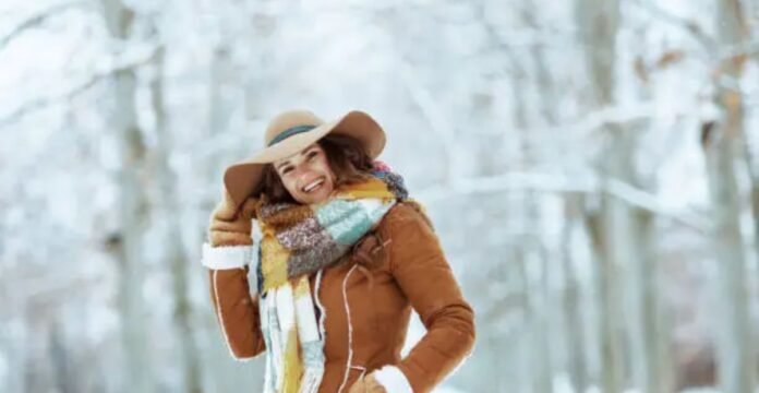 Alt text: A smiling woman in a brown hat, brown jacket, and a colorful scarf stands in a snowy forest with snow-covered trees in the background. Sustainable Wool Winter Coat: