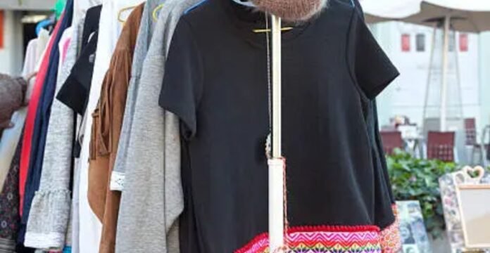 Alt text: A rack of clothes at an outdoor market. Items include a black dress with colorful embroidery at the hem, a brown fringed jacket, and a grey sweater. Market tables with umbrellas and chairs are blurred in the background. Recycled Fabric Winter Coat: