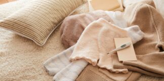 Blankets and Bedding Fabric Guide for Comfort and Warmth Up A cozy scene showing folded blankets and towels on a fluffy beige rug. A striped pillow, a small white thermometer or moisture tester on a card, and a beige knit sweater are arranged among the textiles. Blankets and Bedding fabic