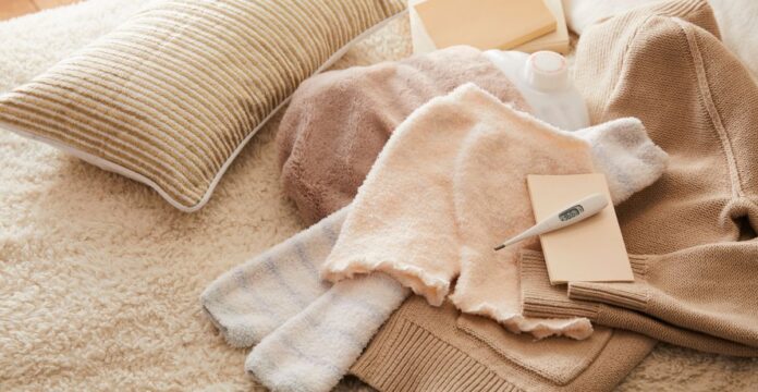 A cozy scene showing folded blankets and towels on a fluffy beige rug. A striped pillow, a small white thermometer or moisture tester on a card, and a beige knit sweater are arranged among the textiles. Blankets and Bedding fabic